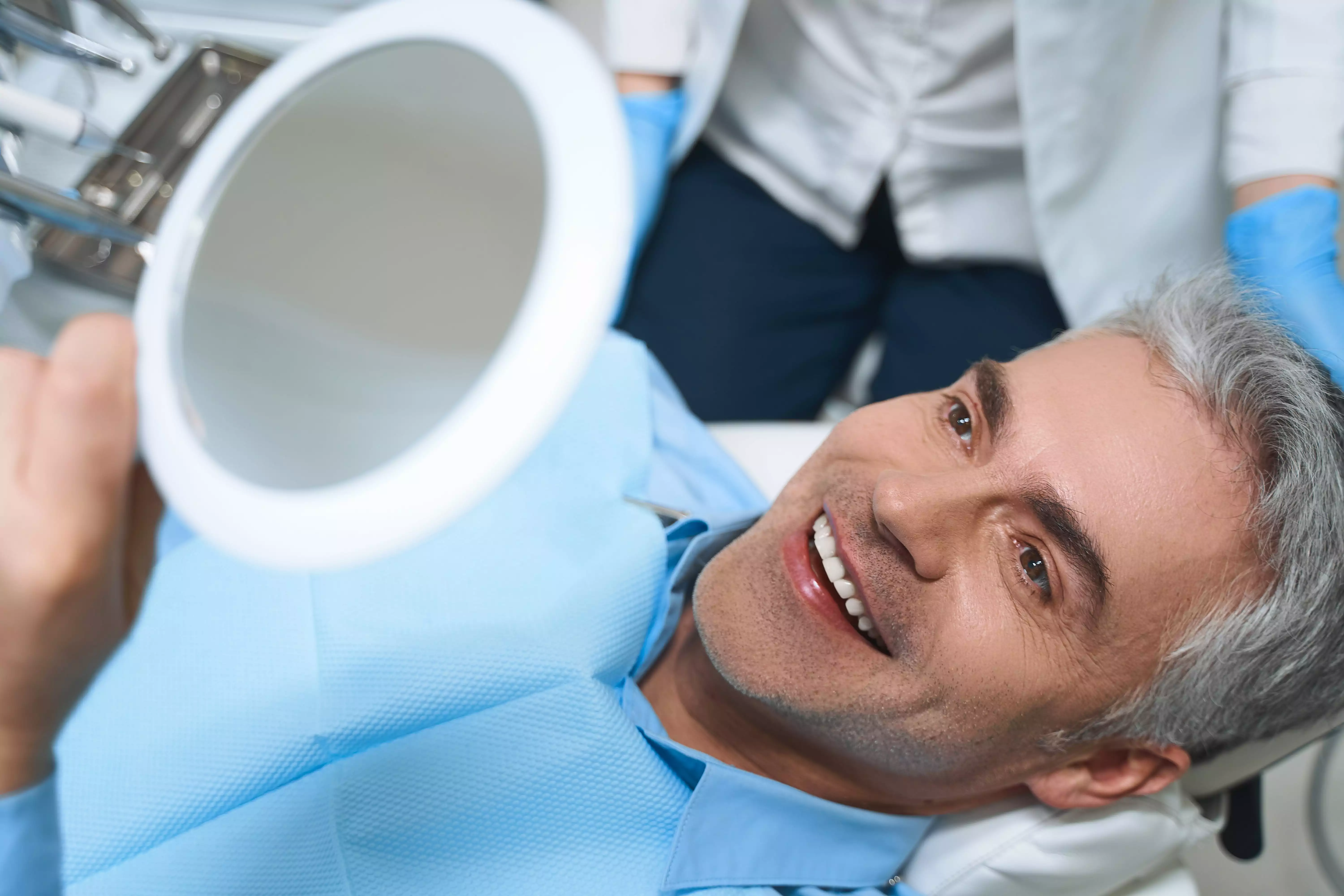 joyful-male-is-lying-chair-looking-into-mirror-while-being-delighted-with-dentist-work-min